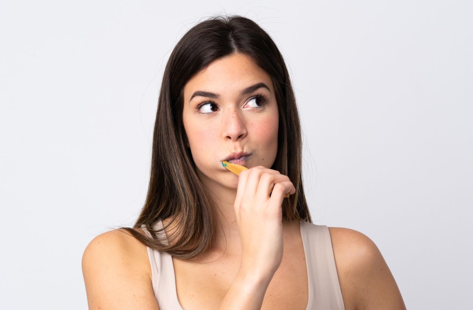 A woman brushing her teeth and looking up to the right with a thoughtful expression