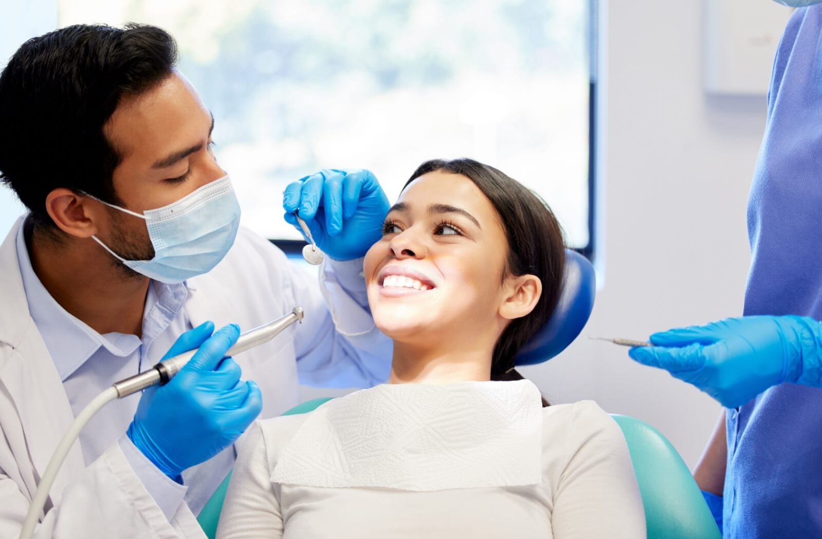 Patient smiling at dentist during teeth cleaning with dental hygienist standing nearby.