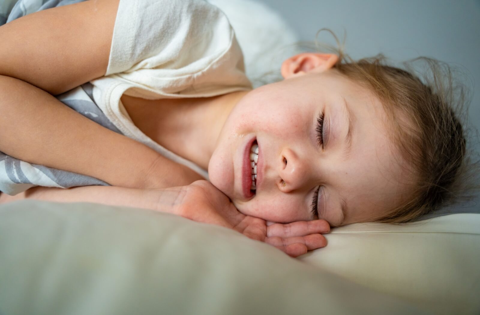 Close-up of a young child peacefully sleeping on their side with a slight smile, showing their teeth while resting on a pillow.